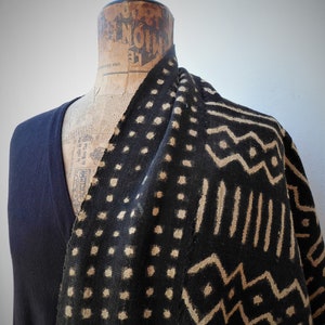 Peut inclure: Une écharpe en tissu à motifs noirs et beiges avec un design géométrique. L'écharpe est drapé sur les épaules d'un mannequin.