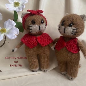 Puede incluir: Dos muñecos de gato tejidos. Un muñeco de gato lleva una diadema roja y un chaleco rojo. El otro muñeco de gato lleva un chaleco rojo. Los muñecos son marrones y beige con detalles faciales negros. El texto "KNITTING PATTERN PDF EN/ES/FR" es visible.