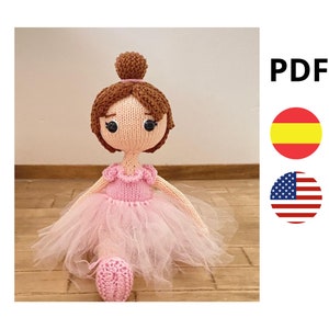 Puede incluir: Muñeca bailarina de punto con pelo castaño, vestido rosa y tutú de tul. La muñeca está sentada sobre una superficie de madera.
