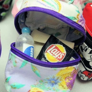 May include: A purple and white floral fabric pouch with a zipper closure. The pouch is open and contains a small clear bottle, a small bag of Lay's potato chips, and a small amount of white fabric.