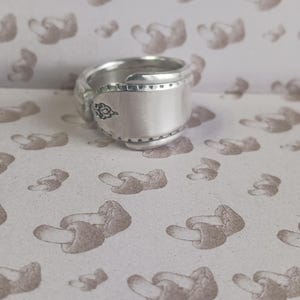 May include: A silver ring crafted from a spoon, featuring a decorative design on the side. The ring has a wide band and a unique, repurposed aesthetic. The background is a pattern of mushroom illustrations.