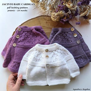 Pode incluir: Três cardigans de bebé de malha, em tons de branco e roxo, com fechos de botões. Os cardigans são exibidos numa superfície de madeira, com flores secas no fundo. O texto "JACINTO BABY CARDIGAN" e "preemie - 24 months" é visível.