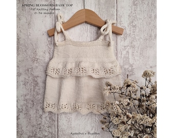 May include: A white knitted baby top with a ruffled trim and tie straps. The top is hanging on a wooden hanger against a white wood background. The text "SPRING BLOSSOMS BABY TOP" and "Pdf Knitting Pattern 0-36 months" is visible on the image. The text "Agasalhos e Bugalhos" is visible at the bottom of the image.
