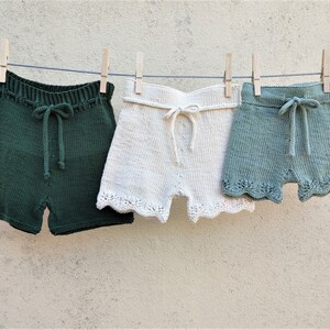 May include: Three pairs of knitted shorts hanging on a clothesline. The shorts are green, white, and light blue. The shorts have a scalloped edge.