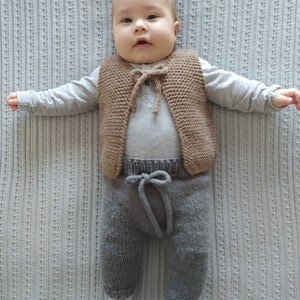 May include: A baby wearing a brown knitted vest with a tie closure and gray knitted pants. The baby is lying on a white blanket.
