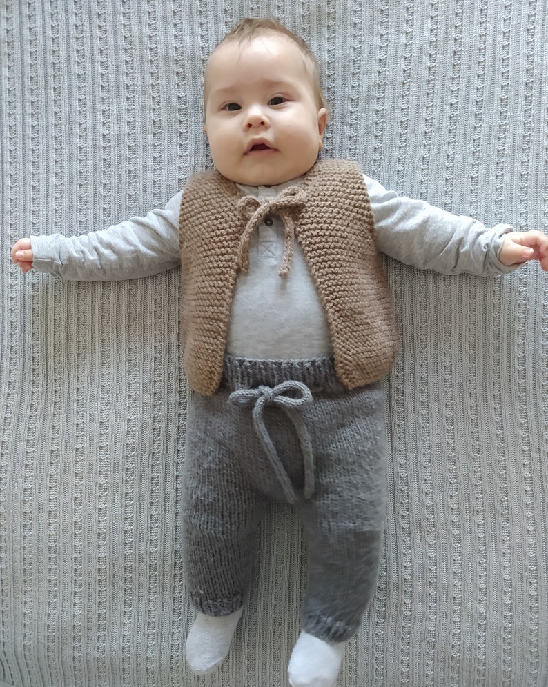 May include: A baby wearing a brown knitted vest with a tie closure and gray knitted pants. The baby is lying on a white blanket.