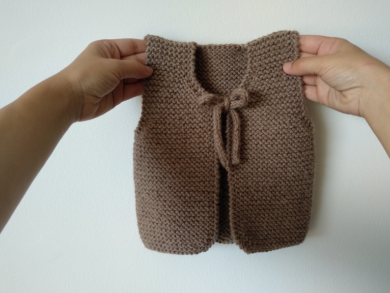 May include: A brown knitted vest with a tie closure. The vest is sleeveless and has a simple design.