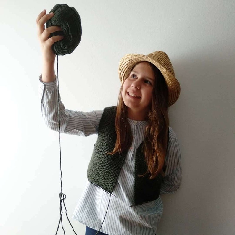 May include: A young person wearing a straw hat and a green knitted vest holds a ball of dark green yarn. The yarn is attached to a string.