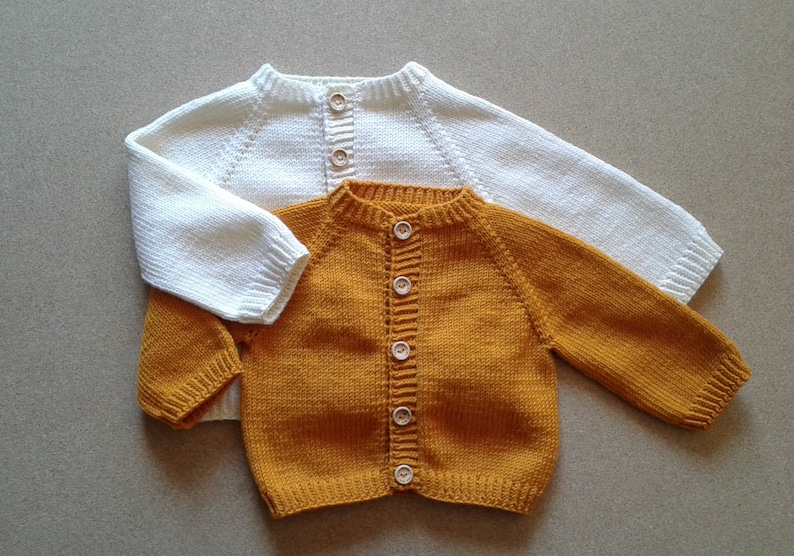 May include: Two knitted baby cardigans, one white and one mustard yellow, with wooden buttons.