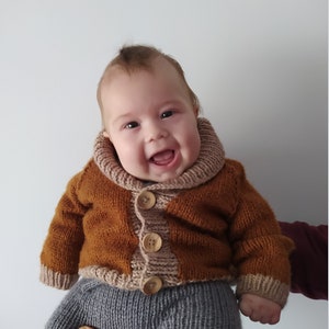 May include: A hand-knitted baby cardigan in a rich tobacco brown colour featuring a wide, ribbed beige shawl collar and cuffs. The garment is fastened with three large, round, light-coloured wooden buttons down the centre front, paired with grey knit trousers.