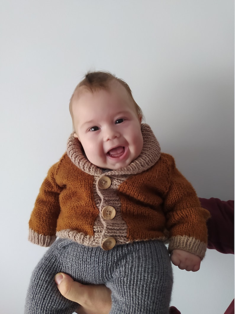 May include: A hand-knitted baby cardigan in a rich tobacco brown colour featuring a wide, ribbed beige shawl collar and cuffs. The garment is fastened with three large, round, light-coloured wooden buttons down the centre front, paired with grey knit trousers.