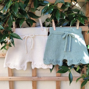 May include: Two pairs of crocheted shorts, one white and one light blue, hanging on a wooden fence. Both pairs have a scalloped edge and a tied bow at the waist.