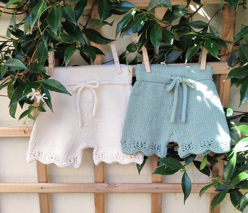May include: Two pairs of crocheted shorts, one white and one light blue, hanging on a wooden fence. Both pairs have a scalloped edge and a tied bow at the waist.