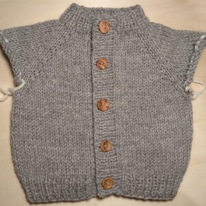 Puede incluir: Un c&aacute;rdigan de punto gris sin mangas con cuello y dobladillo acanalados, una tapeta frontal con cinco botones redondos de madera y bucles de hilo blanco que sobresalen de las sisas en los lados izquierdo y derecho.