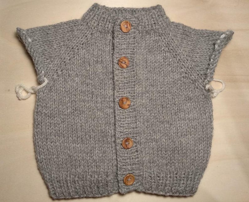Puede incluir: Un c&aacute;rdigan de punto gris sin mangas con cuello y dobladillo acanalados, una tapeta frontal con cinco botones redondos de madera y bucles de hilo blanco que sobresalen de las sisas en los lados izquierdo y derecho.