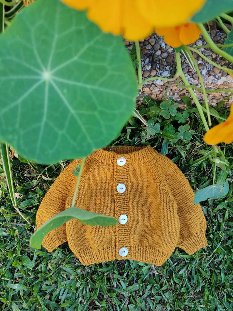 May include: A mustard yellow knitted cardigan with a button closure. The cardigan is lying on a bed of green grass.