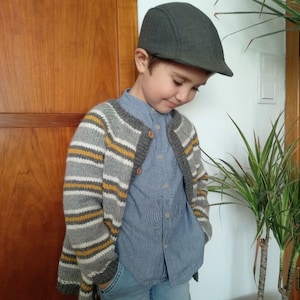 Puede incluir: Una gorra plana gris carb&oacute;n, un c&aacute;rdigan de punto de manga larga con rayas horizontales en gris, blanco y amarillo mostaza con botones de madera, una camisa azul claro de botones con cuello peque&ntilde;o y pantalones vaqueros.