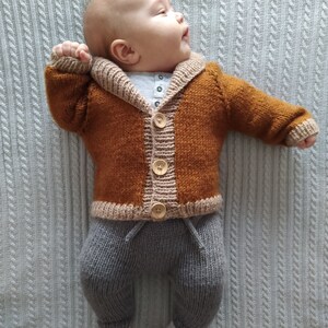 May include: A knitted baby outfit consisting of a long-sleeved, rust-coloured cardigan with a beige shawl collar, cuffs, and hem, fastened with three light-coloured wooden buttons. The outfit includes grey knit trousers with a drawstring waist.
