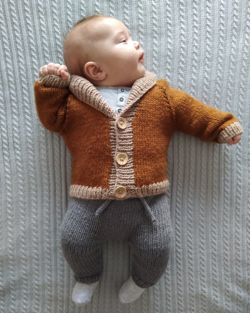 May include: A knitted baby outfit consisting of a long-sleeved, rust-coloured cardigan with a beige shawl collar, cuffs, and hem, fastened with three light-coloured wooden buttons. The outfit includes grey knit trousers with a drawstring waist.