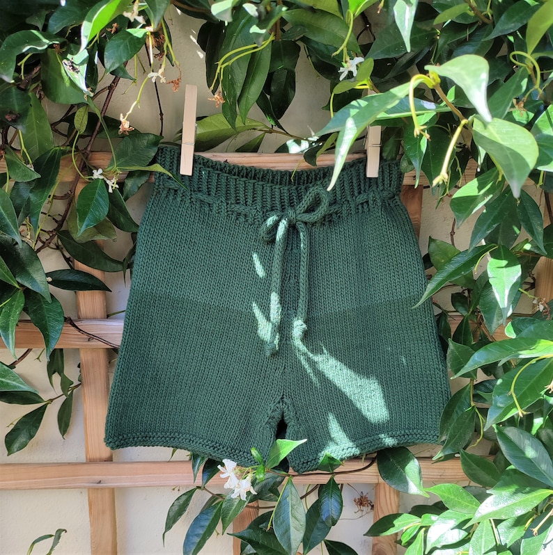 May include: Dark green knitted shorts with a drawstring waist. The shorts are hanging on a wooden trellis with green vines.