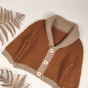 May include: A knitted cardigan in a rich rust-brown colour featuring a contrasting beige shawl collar, cuffs, and hem. The garment is secured with three light-coloured wooden buttons down the front, showcasing a classic chunky knit texture.