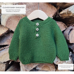 May include: A green knitted baby sweater with a button fastening. The sweater is hanging on a wooden hanger against a background of stacked logs of wood. The text "CLOVER BABY SWEATER" and "PDF KNITTING PATTERN PREEMIE-24 MONTHS" is printed above the sweater. The text "AGASALHOS E BUGALHOS BY SANDRA MAGALHÃES" is printed below the sweater.