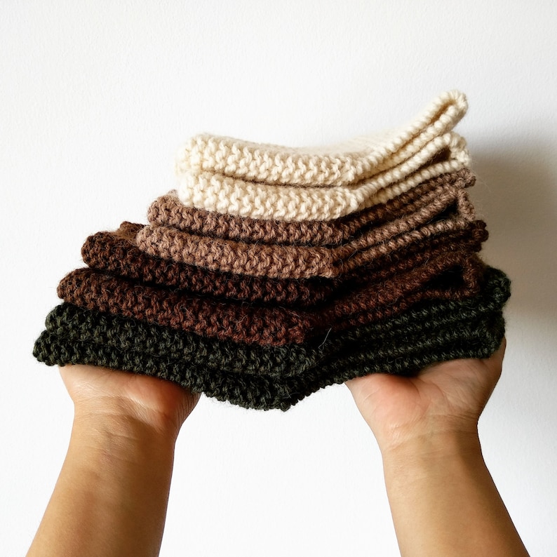 May include: A stack of five knitted squares in various shades of brown and green. The squares are folded and stacked on top of each other, with the green square on the bottom and the white square on top.