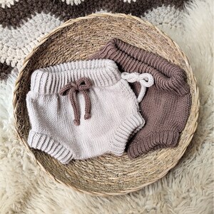 May include: Two pairs of knitted baby bloomers in a wicker basket. One pair is beige and the other is brown.