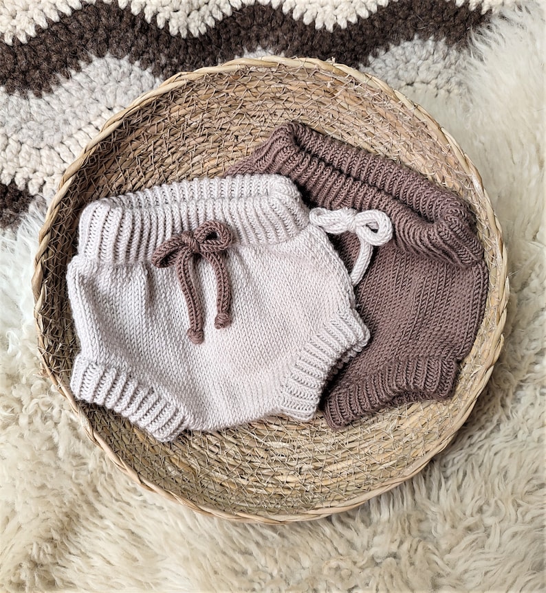 May include: Two pairs of knitted baby bloomers in a wicker basket. One pair is beige and the other is brown.