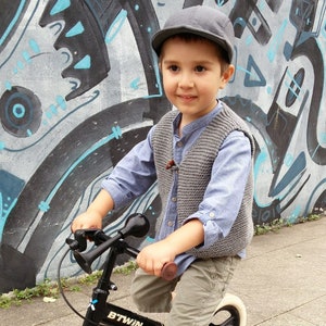 May include: A young boy wearing a gray knitted vest and a blue shirt rides a black and silver bicycle with a horn. The bicycle has a black seat and a white front wheel. The boy is wearing a gray cap and is smiling.