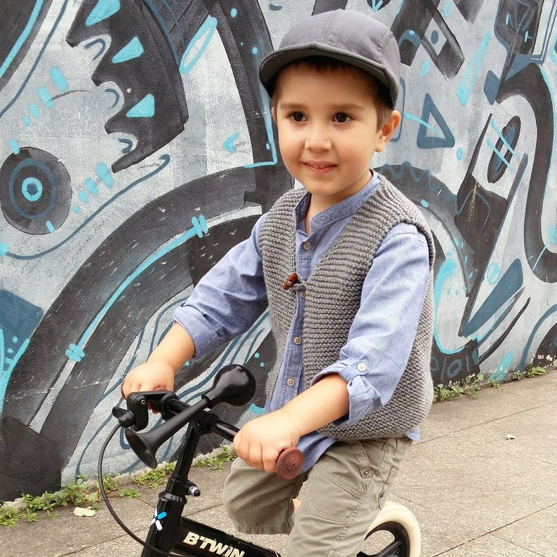 May include: A young boy wearing a gray knitted vest and a blue shirt rides a black and silver bicycle with a horn. The bicycle has a black seat and a white front wheel. The boy is wearing a gray cap and is smiling.