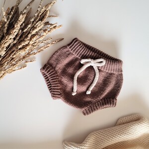 May include: Brown knitted baby bloomers with a white drawstring tie. The bloomers are made of a soft, comfortable material and have a relaxed fit.
