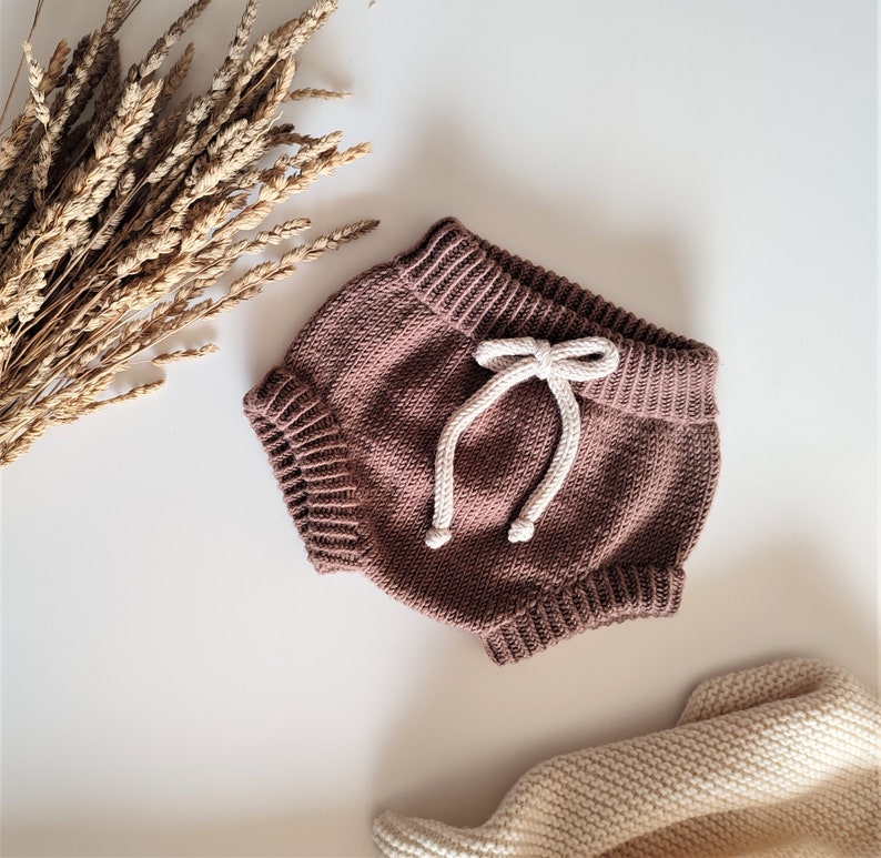 May include: Brown knitted baby bloomers with a white drawstring tie. The bloomers are made of a soft, comfortable material and have a relaxed fit.