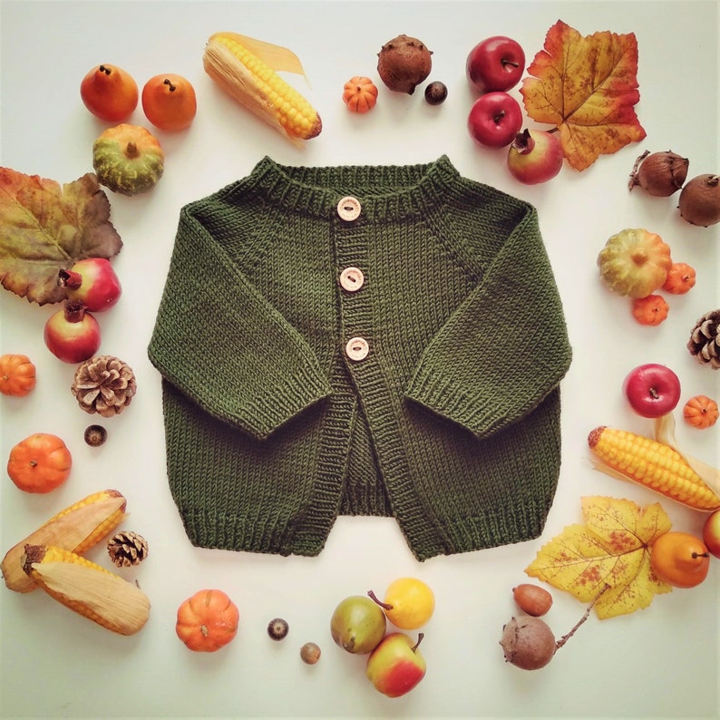 May include: A dark green knitted cardigan with a button-up front. The cardigan is laid out on a white surface with autumnal decorations around it.