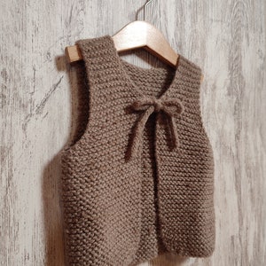 May include: A brown knitted vest with a tie closure. The vest is hanging on a wooden hanger against a white wood background.