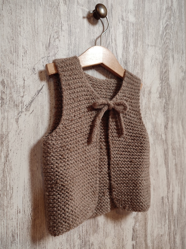May include: A brown knitted vest with a tie closure. The vest is hanging on a wooden hanger against a white wood background.