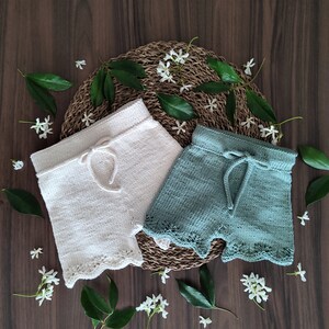 May include: Two pairs of knitted baby shorts, one white and one green, with a lace trim. The shorts are laid out on a woven mat with white flowers.