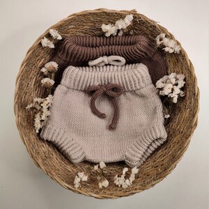 May include: A pair of knitted baby bloomers in a light beige color with a brown knitted waistband and bow. The bloomers are displayed in a woven basket with dried flowers.