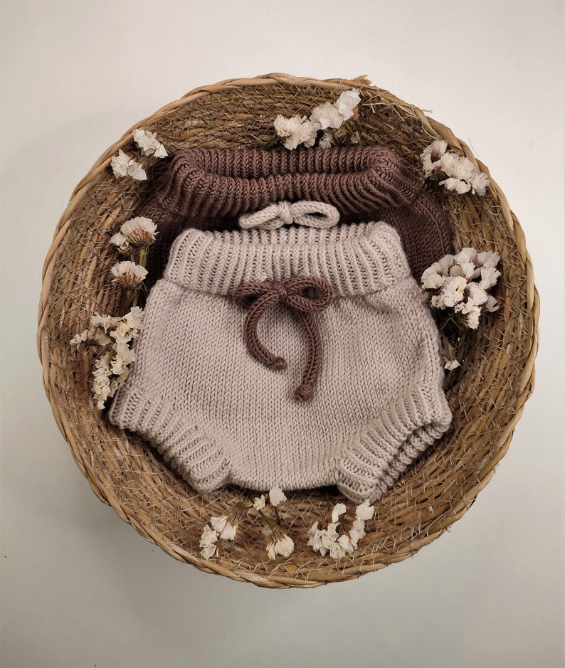 May include: A pair of knitted baby bloomers in a light beige color with a brown knitted waistband and bow. The bloomers are displayed in a woven basket with dried flowers.