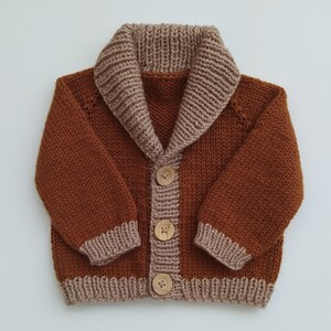May include: A long-sleeved knit cardigan in a deep rust-brown colour featuring a contrasting light tan ribbed shawl collar, cuffs, and hem. The front closure includes three round, light-coloured wooden buttons with four holes each.