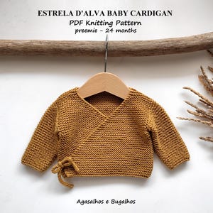 Könnte beinhalten: Senfgelbe Baby-Strickjacke mit langen Ärmeln, die an einem Holzkleiderbügel hängt. Der Wickelpullover hat einen Kordelverschluss an der Seite. Der Text "ESTRELA D'ALVA BABY CARDIGAN" steht oben. Das PDF-Strickmuster ist für Frühchen bis 24 Monate.