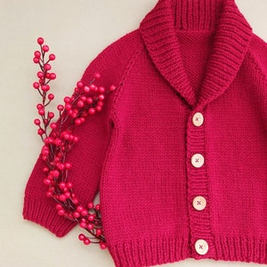 May include: A vibrant magenta knitted cardigan featuring a wide shawl collar, long sleeves, and four light-coloured wooden buttons down the front. A decorative branch with small, round, bright red berries is positioned along the left side.