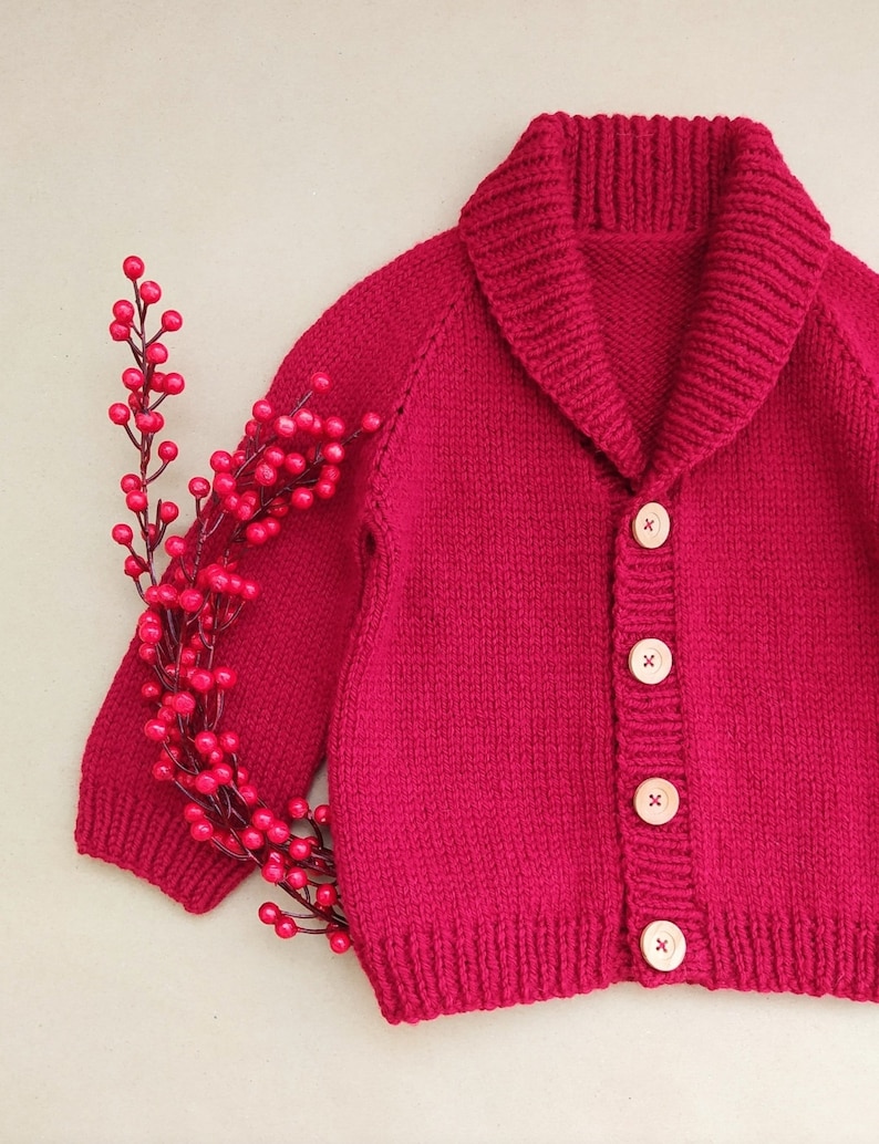 May include: A vibrant magenta knitted cardigan featuring a wide shawl collar, long sleeves, and four light-coloured wooden buttons down the front. A decorative branch with small, round, bright red berries is positioned along the left side.
