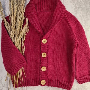 May include: A deep magenta knit cardigan featuring a wide shawl collar, long sleeves, and four light-coloured wooden buttons down the front. The garment is displayed alongside a bundle of dried wheat stalks. The text Agasalhos e Bugalhos is visible.