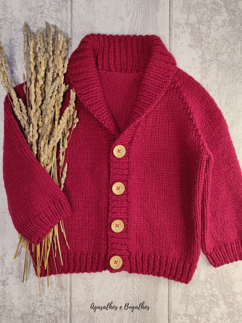 May include: A deep magenta knit cardigan featuring a wide shawl collar, long sleeves, and four light-coloured wooden buttons down the front. The garment is displayed alongside a bundle of dried wheat stalks. The text Agasalhos e Bugalhos is visible.