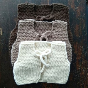 May include: Three knitted vests in different shades of brown and white. The vests are sleeveless and have a tie at the front.