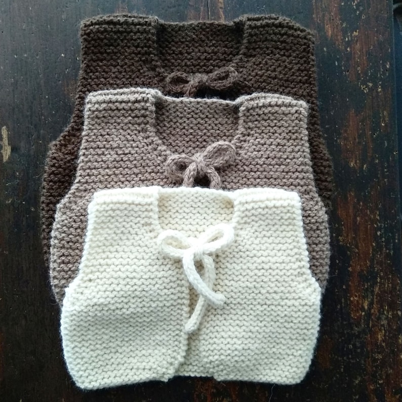May include: Three knitted vests in different shades of brown and white. The vests are sleeveless and have a tie at the front.