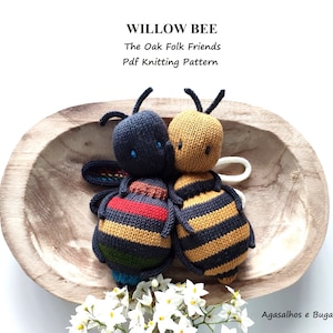 May include: Two knitted bee toys, one black and yellow with red stripes, the other yellow and black, sitting in a wooden bowl. The bowl is surrounded by white flowers. The text "Willow Bee The Oak Folk Friends Pdf Knitting Pattern" is above the bees.