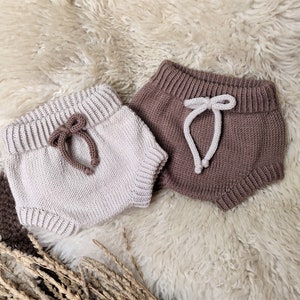 May include: Two pairs of knitted baby bloomers. One pair is beige with brown ties and the other pair is brown with white ties.