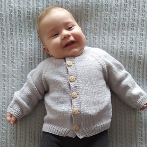 May include: A baby wearing a light purple knitted cardigan with wooden buttons. The cardigan has a ribbed texture and a classic button-up design.